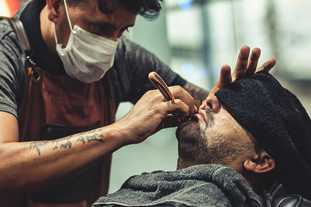 A masked barber in an apron shaves a client’s beard with a straight razor as the client reclines with a dark towel over his eyes